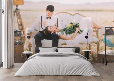 The happy groom with the wedding bouquet is lying on the groom`s laps and he is petting her face at the background of the field. Wall mural